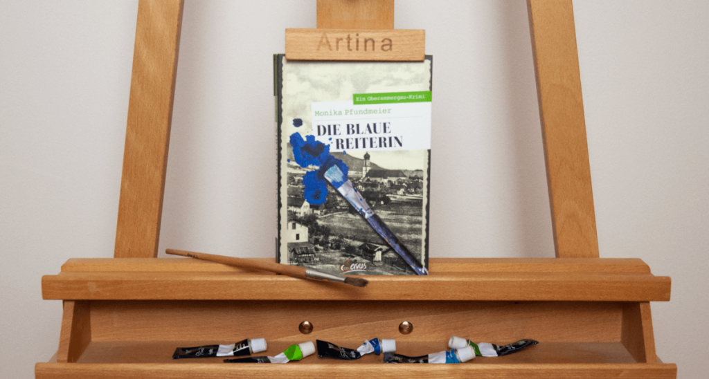 The picture shows the book "Die blaue Reiterin" by Monika Pfundmeier standing on an easel. A paintbrush stands next to it and below the book lie 5 paint tubes in black, dark blue, bright blue, dark green and bright green.