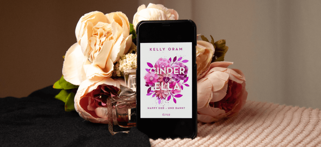 An iPhone screen shows the book cover of "Happily Ever After" by Kelly Oram is showing. The iPhone stands in front of a fake flower bouquet and hides half of a bottle of Miss Dior perfume