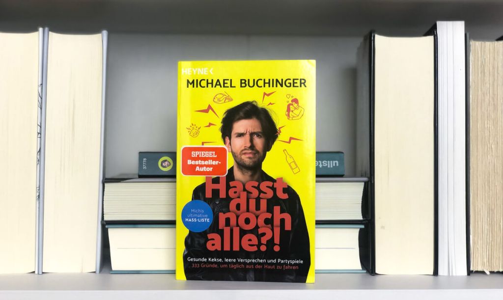 Michael Buchinger's Buch "Hasst du noch alle?!" steht auf einem weißen Bücherregal. Alle anderen Buchrücken in diesem Regal sind weggedreht, sodass nur deren Seiten zu sehen sind und man sich auf "Hasst du noch alle?!" konzentrieren kann.