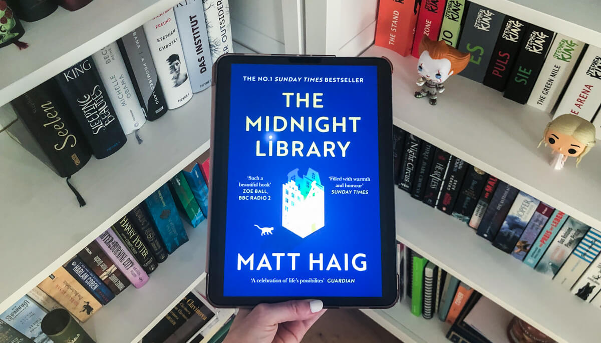 The cover of "The Midnight Library" by Matt Haig is shown on an iPad. The iPad is held with one hand while in the background you can see two bookshelves.